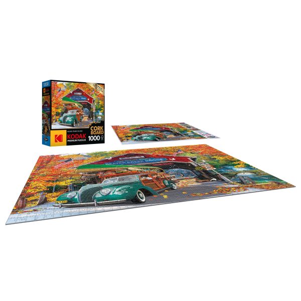 A completed RoseArt - Kodak Cork - Covered Bridge - 1000 Piece Jigsaw Puzzle depicting a vintage car scene is displayed flat on a surface, with the puzzle box standing nearby showing the same image.
